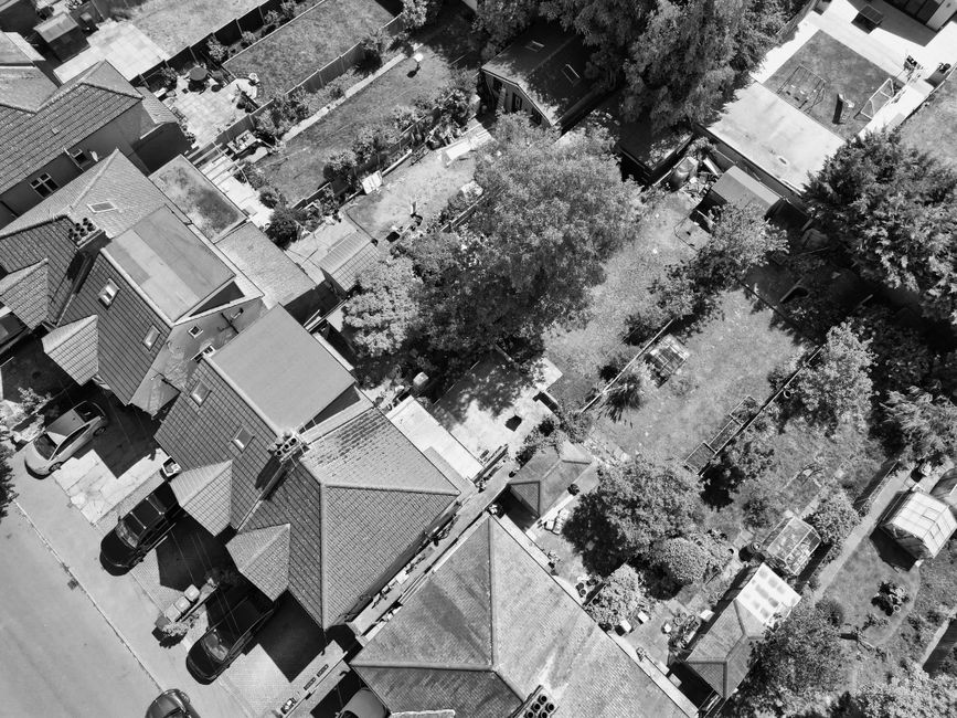 An aerial black-and-white view of a residential garden and surrounding houses, showcasing a well-maintained backyard with a mix of grassy lawn areas, garden beds, and paved pathways. Several mature tr