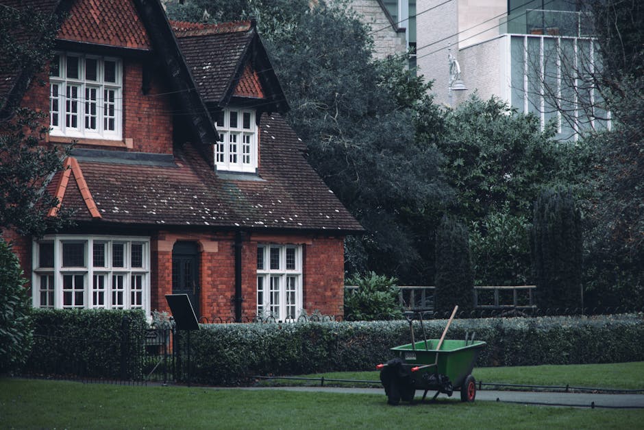 A traditional red-brick house with a steeply pitched tiled roof and multiple white-framed casement windows, set within a well-maintained garden. The garden features a neatly trimmed hedge bordering a 